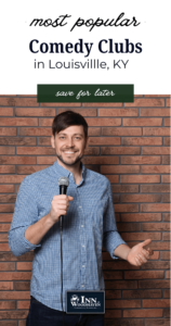 White comedian wearing a blue shirt holds a microphone and smiles in front of a red brick wall