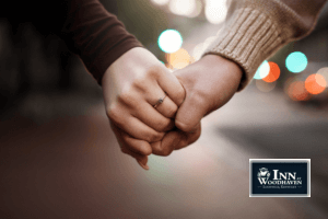 Two hands, one with an engagement ring, are clasped in front of a multicolored blurred background.