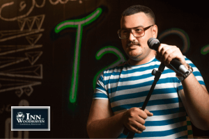 White male comedian in a blue and white striped shirt holds a microphone in front of a black background