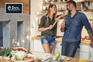 Bearded man in a denim top and jeans holds a beer in a kitchen as he smiles at a blonde woman in a green shirt and jean shorts, standing in a kitchen.