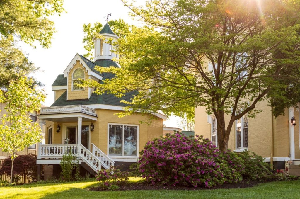 View from front yard looking at the Rose Cottage, a yellow octagonal structure that is painted yellow with a green roof. A large tree visible in the foreground.