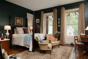 Spacious room with dark evergreen painted walls and two gothic style windows. A queen four-post bed in the center of the room with a white bedspread and decorative pillows. Two chairs sit at the foot of the bed on a large decorative area rug.