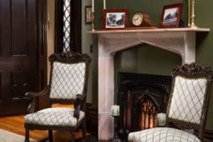 Antique chairs in front of a lit fireplace and old pictures on the mantle.