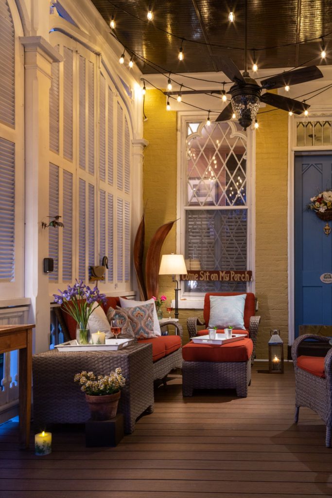 Back patio lit with Edison lights. Hellow walls with white shutters and a blue door with floral arrangement hanging on it. Red upholstered patio furniture and assorted drinks implying friends hanging out.