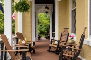 Sunny front porch with rocking and adirondack chairs. A hanging plant and a vase of pink flowers and two glasses of iced tea with lemon complete this summer themed picture.