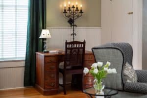 Wood desk with a dark wood chair. White lamp on the desk with a crystal vase of white flowers in front.