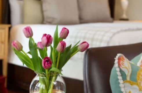 Purple tulips on a table next to a love seat with a book and glasses next to it. A bed and lamp in the background