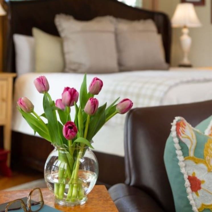 Purple tulips on a table next to a love seat with a book and glasses next to it. A bed and lamp in the background