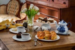 set table with ornate teapot, vase of flowers, croissant sandwiches, tea and cookie selection