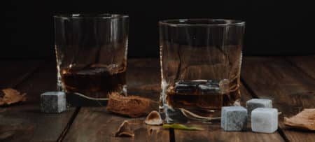 Two rocks glasses with golden bourbon whiskey on a wooden table covered in natural materials and four grey whiskey stones.